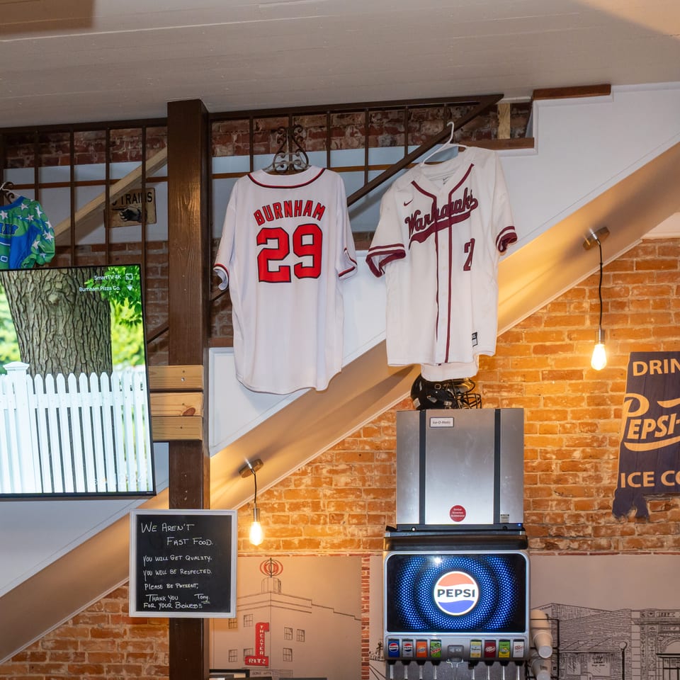 Burnham Pizza restaurant interior featuring hanging sports jerseys, exposed brick walls, Pepsi fountain, television, and staircase.