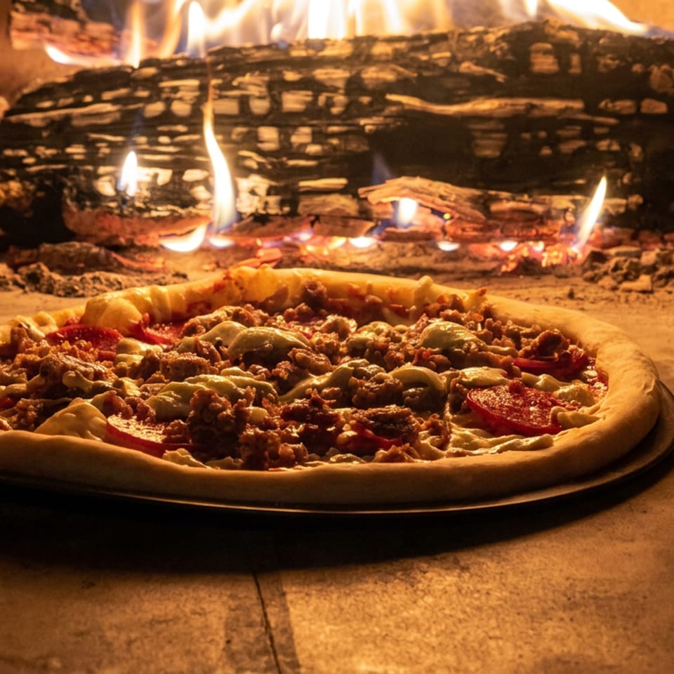 Pepperoni, sausage, and mushroom pizza baking in a wood-fired oven with glowing logs and flames behind it.