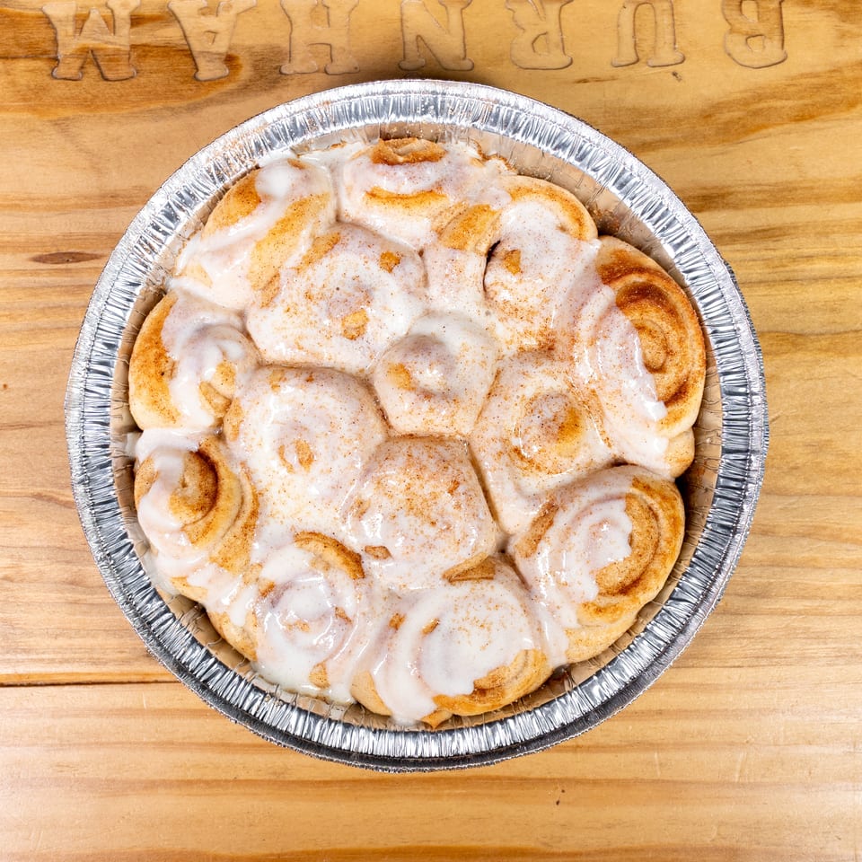 Freshly baked cinnamon rolls covered in white icing, arranged in a round aluminum pan on a wooden table.