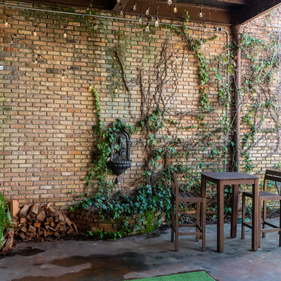 Rustic restaurant patio with ivy-covered brick walls, string lights, a fountain, ferns, stacked firewood, and a tall wooden table.
