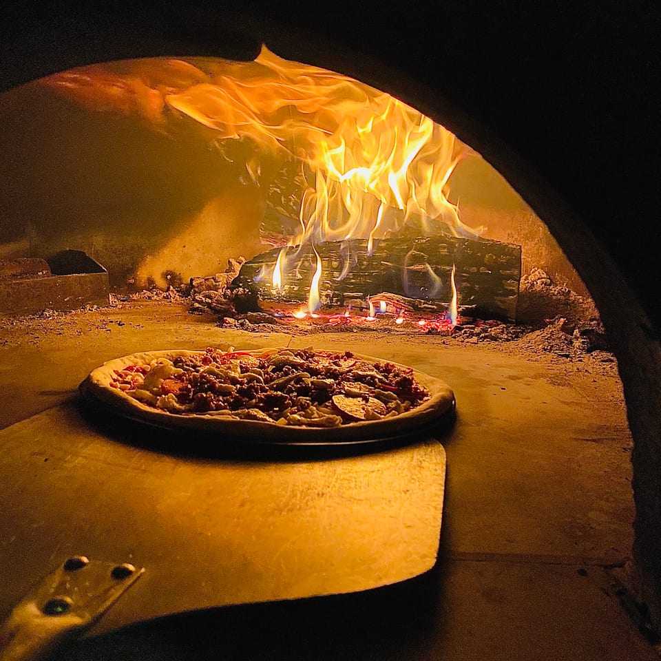 Topped pizza baking in a wood-fired oven with bright flames and glowing embers.