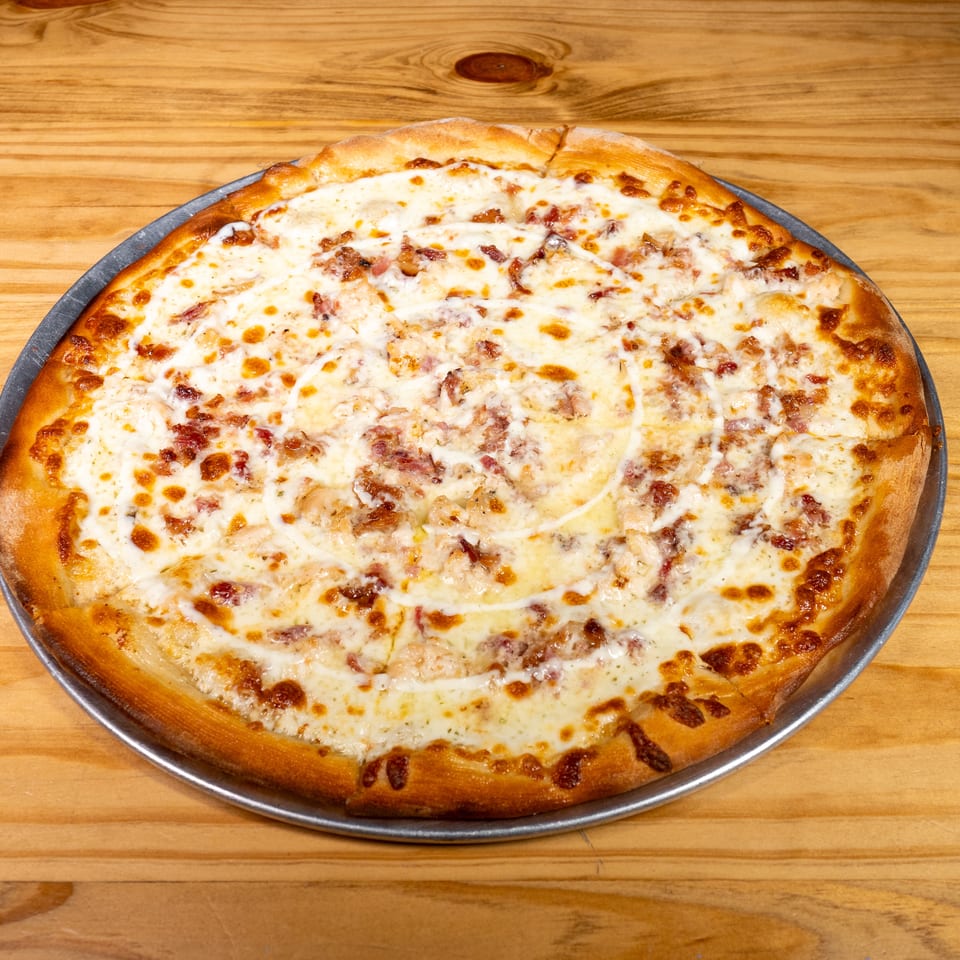 Large round pizza topped with melted cheese and bacon pieces on a metal pan atop a wooden table.