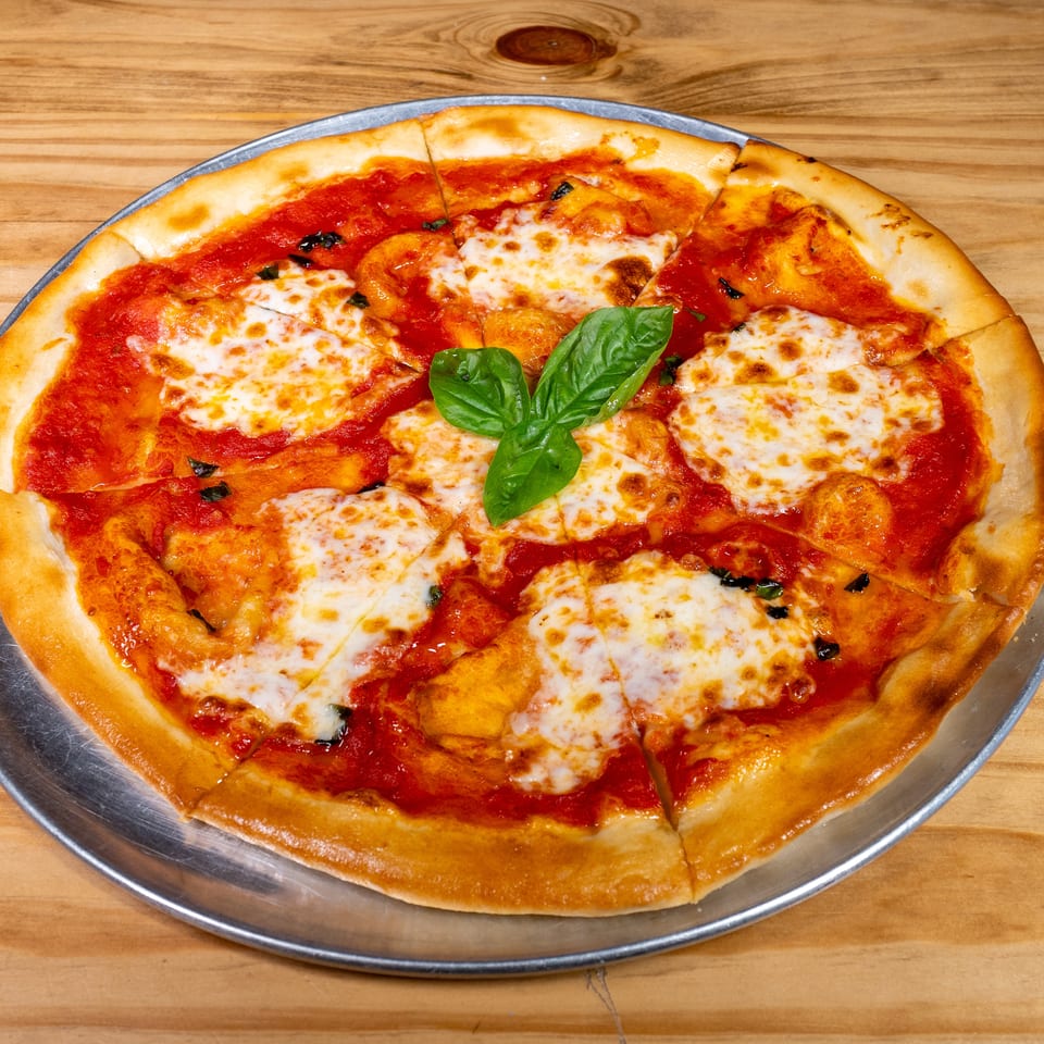 Large cheese pizza with tomato sauce, basil leaves, and browned toppings on a metal tray atop a wooden table.