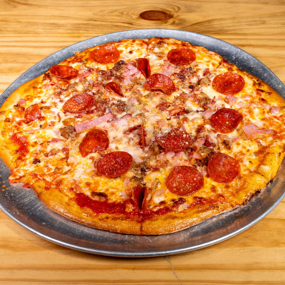 Large meat-lover’s pizza topped with pepperoni, ham, sausage, and melted cheese on a metal tray atop a wooden table.