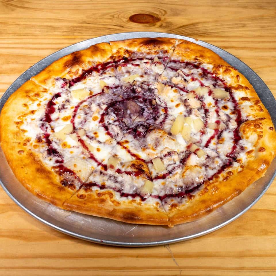 Cheesy pizza topped with pineapple, meat, and red berry sauce on a metal tray over a wooden table.