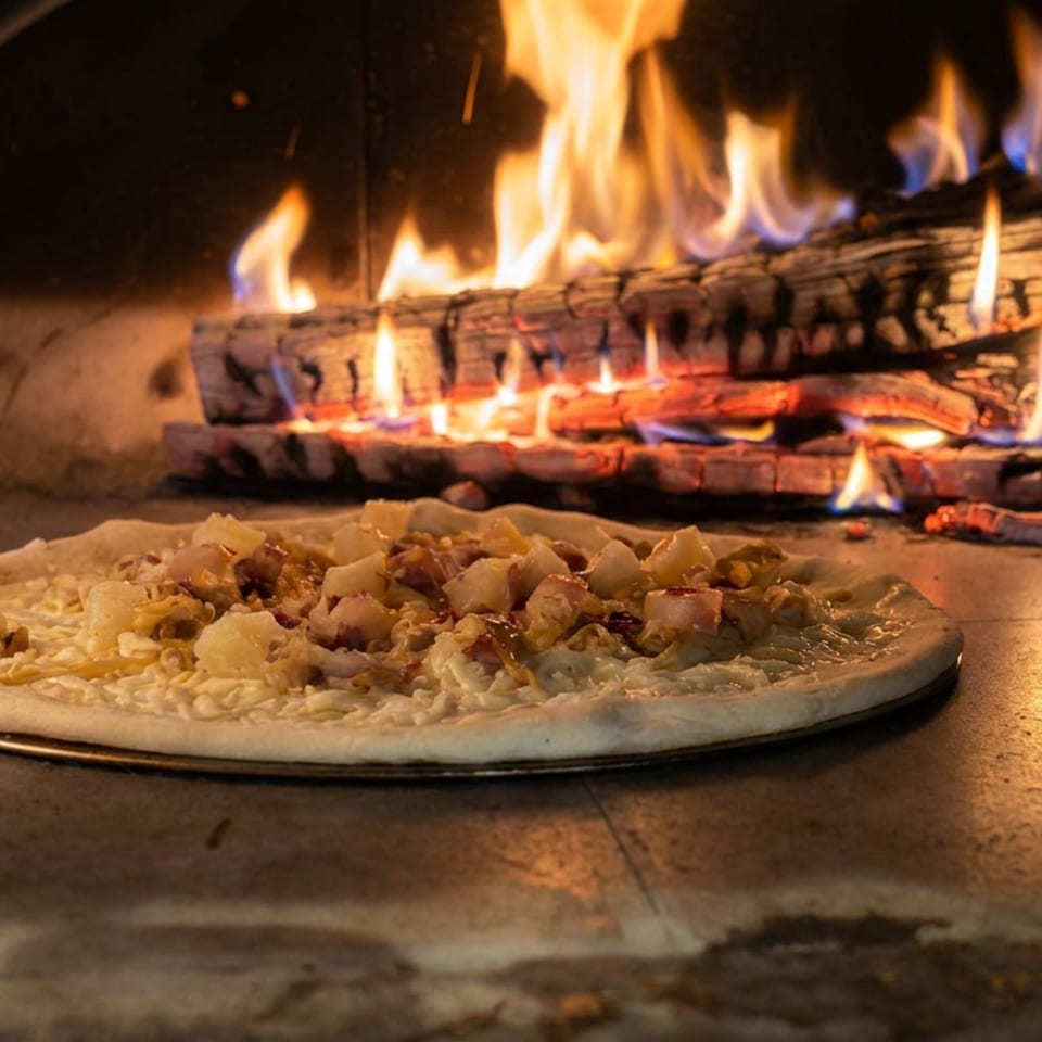 Unbaked topped pizza rests on a metal tray inside a wood-fired oven, with flames and burning logs behind it.