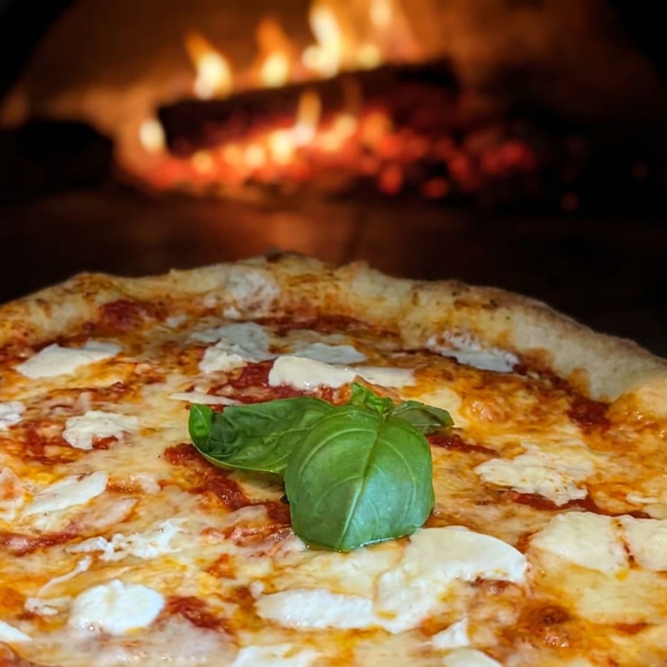 Freshly baked Margherita pizza with melted cheese, tomato sauce, and basil in front of a wood-fired oven.