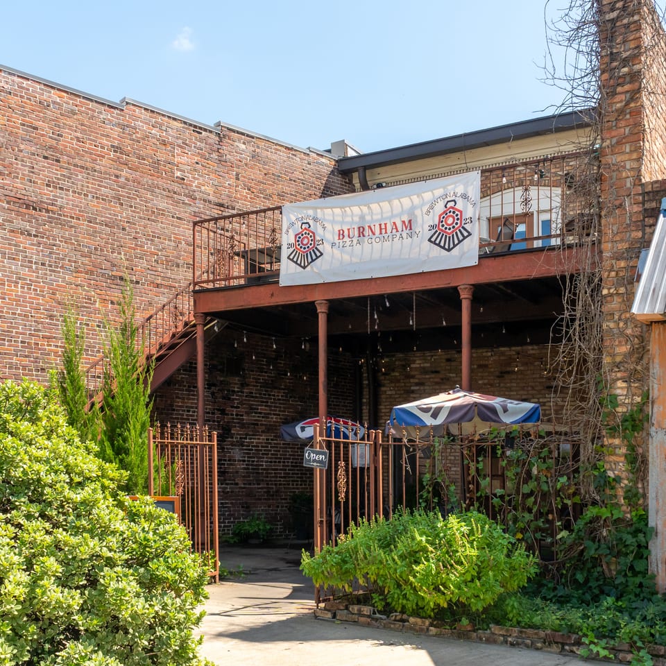Burnham Pizza Company entrance in a brick courtyard with patio seating, greenery, stairs, and an open sign.
