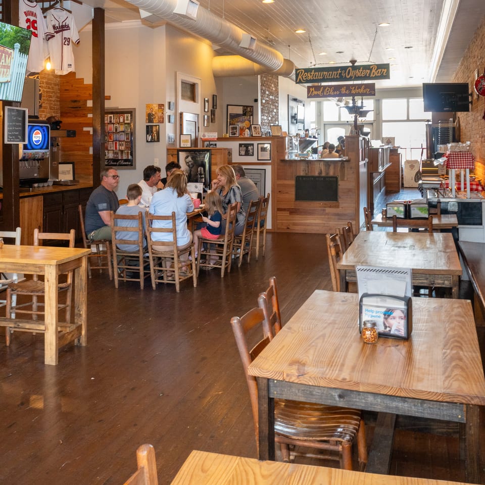 Rustic restaurant and oyster bar interior with exposed brick walls, wooden tables, sports memorabilia, and diners seated near the counter.
